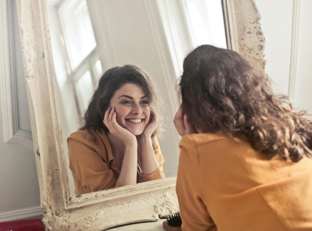 pexels-photo-774866-774866 A cheerful woman smiles at her reflection in a vintage-style mirror, exuding positivity and warmth.
